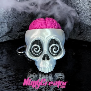 May include: A skull-shaped keychain with a pink brain inside. The skull is white with black spiral eyes and a silver key ring. The text "NiftyCreator.com" is visible at the bottom. The background is black with a smoky effect.