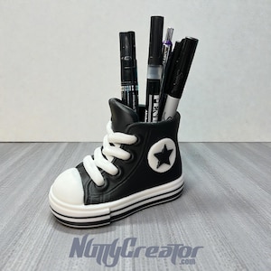 May include: A black and white sneaker-shaped desk organizer holding several black pens and markers. The organizer has white laces, a white toe cap, and a white star emblem on the side. The base has a white stripe.