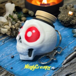 May include: A white skull-shaped keychain with large red eyes and a tan top. The keychain has a silver clasp and the text "NuttyCreator.com" in yellow. The design is detailed and has a Halloween or gothic theme.