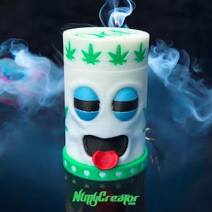 May include: A white candle with a cartoon face design, featuring blue eyes, a black mouth, and a red tongue. The top has green cannabis leaf accents. The base has a green band. The candle is lit, with smoke in the background. The text "NuttyCreator.com" is at the bottom.
