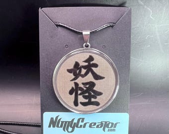 May include: A round pendant necklace with a black cord. The pendant has a silver-colored frame and a tan center with black Japanese characters. The necklace is displayed on a black card with a blue sticker that reads "NiftyCreator.com".