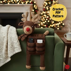 Può includere: Un organizer a forma di renna all'uncinetto marrone fatto a mano con due tasche, un naso rosso e corna. L'organizer è progettato per essere appeso sul braccio di un divano verde. L'immagine include il testo "Crochet PDF Pattern".