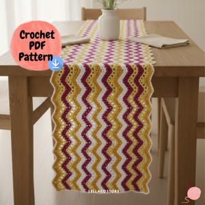 May include: A handmade crochet table runner with a chevron pattern in white, gold, and burgundy. The text "Crochet PDF Pattern" is in a pink circle in the upper left corner. The table runner is displayed on a wooden table.