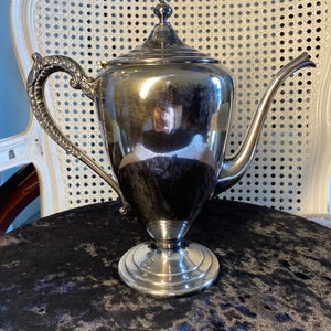 May include: A silver teapot with a decorative handle and a polished finish. The teapot is sitting on a black velvet surface.