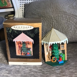 May include: A white and brown Christmas tree ornament shaped like a gazebo with a green tree inside. The ornament has a white roof with icicles and a Santa Claus figure climbing the stairs. The box reads "Keepsake Ornament, Treasured Memories Light, Magic".