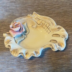 Könnte beinhalten: Dekorative, cremefarbene Keramikschale mit gewelltem Rand, verziert mit goldfarbenen Verzierungen. Eine rosa und lavendelfarbene Rose mit grünen Blättern ziert die Seite. Die Schale hat eine strukturierte Oberfläche und dient wahrscheinlich zur Präsentation.