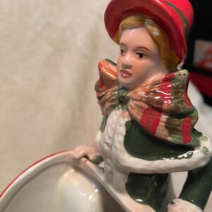May include: A ceramic figurine of a person wearing a red hat with a green band, a green and white coat, and a red and green scarf. The figurine is holding a white bowl with a red rim.