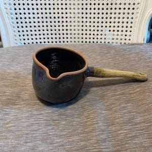 May include: A small, dark brown ceramic pitcher with a light brown handle. The pitcher has a rounded body and a small spout.