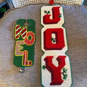 以下が含まれることがあります： 2つのクリスマス飾り。緑色の「NOEL」ドアハンガーには銀色のベルと赤、白、緑の模様があり、「JOY」のサインは赤い文字に緑色のアクセントが付いています。どちらもクロスステッチで作られています。