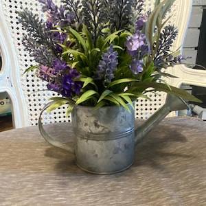 May include: A galvanized metal watering can with a purple and green floral arrangement. The flowers are in a rustic style and the watering can has a distressed finish.