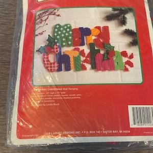 May include: A craft kit for a "Merry Christmas" wall hanging. The design features colourful letters and festive embellishments. The package reads "TEMPTATIONS" at the top and includes instructions and materials for a finished piece approximately 25 cm high and 36 cm wide.