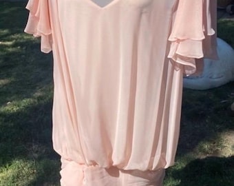 May include: A vintage blush pink dress with a sheer overlay and ruffled sleeves. The dress features delicate lace shoulder embellishments and a draped skirt. The dress is displayed on a mannequin against a backdrop of green grass.