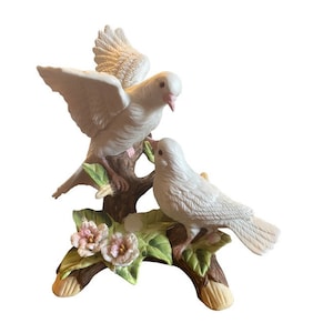 May include: A porcelain figurine of two white doves perched on a branch with green leaves and pink flowers. The doves are in a loving embrace, symbolizing peace and love.