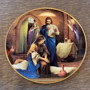 May include: A decorative plate with a gold rim features a religious scene. The image depicts figures in robes, with a central figure in blue and white. The background shows a brick structure and a table with items.