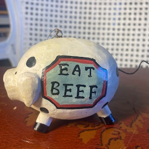 Könnte beinhalten: Weißer Keramik-Schweine-Anhänger mit schwarzen Punkten und einem roten und blauen Schild mit der Aufschrift "EAT BEEF".