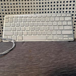 以下が含まれることがあります： QWERTY配列のシルバーとホワイトのワイヤレスキーボード。薄型キー、コマンドキー、オプションキーを備えています。左側から伸びる白い充電ケーブルと、メタリックな仕上げが特徴です。標準的なファンクションキーも配置されています。