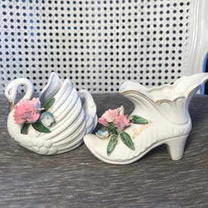Puede incluir: Dos figuritas de porcelana blanca, una con forma de cisne y la otra con forma de zapato. Ambas están decoradas con flores rosas y azules y hojas verdes. El zapato tiene la palabra "Madre" escrita en él.