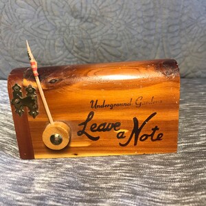 May include: A wooden box with a small American flag sticking out of the top. The box is labeled "Underground Gardens Leave a Note".