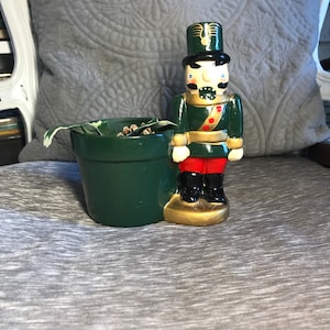 May include: A green ceramic planter with a green and gold nutcracker figurine. The planter has a leafy plant inside. The nutcracker is wearing a red and gold uniform with a black hat.
