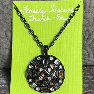 Pode incluir: Um colar de corrente prateada com um pingente redondo com fundo preto e um mosaico de strass transparentes e marrons. O pingente está preso a uma corrente preta. O colar é apresentado em um cartão verde com o texto "Family Treasure Trunk - Etsy".