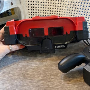 Könnte beinhalten: Ein rot-schwarzes Virtual Boy Gaming-Headset mit einem Doppellinsen-Sichtfenster und einem schwarzen Controller. Das Gerät enthält ein gedrucktes Etikett mit dem Text 1-800-255-3700, Nintendo Authorized Repair Center, und ein schwarzes Kabel.