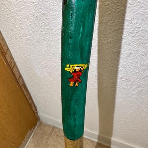 May include: A hand-painted wooden walking stick. The top is painted white and black, transitioning to a vibrant green. A small, colorful figure is depicted on the green section. The bottom portion is a natural wood color.