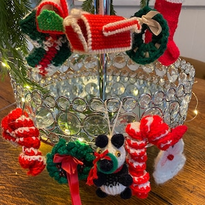 Puede incluir: Una colección de adornos navideños hechos a mano, que incluyen una media, una corona, un bastón de caramelo, un buzón y un oso panda. Los adornos están hechos de hilo y están decorados con colores rojo, blanco y verde.