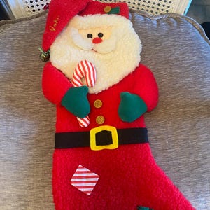 Könnte beinhalten: Ein weihnachtlicher Strumpf aus Plüsch in Form eines Weihnachtsmanns. Der rote Strumpf hat einen weißen Bart, einen roten Hut und einen schwarzen Gürtel mit einer goldenen Schnalle. Der Weihnachtsmann hält eine Zuckerstange. Grüne und gestreifte Flicken verzieren den Strumpf.