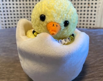 Peut inclure: Un poussin jaune fait à la main avec des yeux noirs et un bec orange, sortant d'un œuf blanc. Le poussin porte un collier de perles avec les lettres "BERT". L'œuf est posé sur une surface marron.