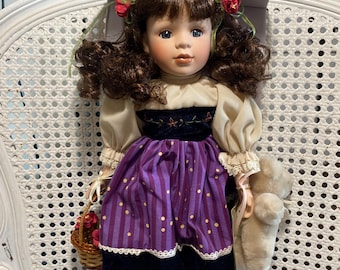 May include: A porcelain doll with brown curly hair, wearing a purple and gold dress with a white blouse and a red flower hat. She is holding a basket of flowers and a teddy bear.