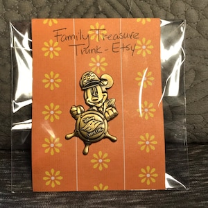 May include: A gold-colored Mickey Mouse pin featuring Mickey dressed as a ship captain. He is holding a steering wheel with the words "Disney Cruise Line" on it. The pin is on a card with a yellow floral pattern and the words "Family Treasure Trunk - Etsy".
