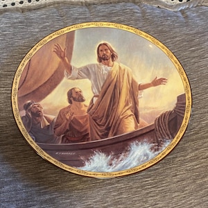 May include: A round porcelain plate with a gold rim features a painting of Jesus Christ walking on water. The scene depicts Jesus with outstretched arms, walking on a stormy sea, with a boat and two disciples in the background. The plate is decorated with a gold border and the word "remarkable" is printed in gold at the bottom.