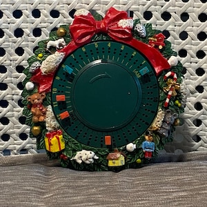 Op de afbeelding: Een groene kerstkrans-timer met een rode strik en verschillende kerstversieringen, waaronder een teddybeer, een huis, een hond, een snoepstok en een speelgoed soldaat. De timer heeft een wijzerplaat met de nummers 1 tot en met 12 en AM/PM-markeringen.