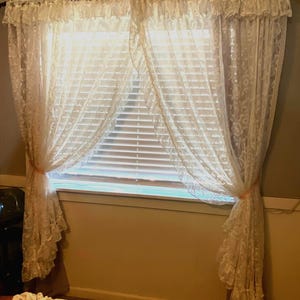 May include: Cream-colored lace curtains framing a window with horizontal blinds. The curtains are tied back with pink ribbons, and the lace has a floral pattern. The window is set in a room with neutral-toned walls.