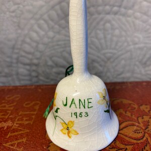 Puede incluir: Campana de cerámica blanca con un esmalte craquelado. La campana está decorada con flores amarillas y hojas verdes. La campana tiene el nombre "JANE" y el año "1953" escrito en ella.