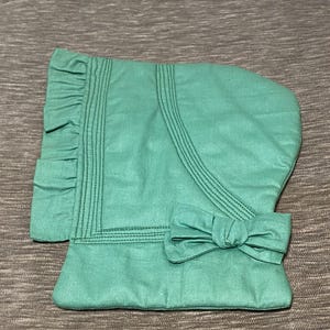 May include: A green fabric bonnet with a ruffle trim and a bow at the back.