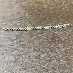 May include: A silver-toned metal link bracelet featuring a repeating pattern of small, rectangular segments connected by rounded, polished links. The bracelet includes a secure clasp mechanism at one end and a matching loop at the other.