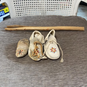 Può includere: Tre paia di mocassini, un paio con perline rosse e bianche, un paio con perline nere e bianche e un paio con un design semplice. I mocassini sono su una superficie di tessuto marrone chiaro.