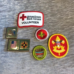 May include: A collection of six embroidered patches, including a red and white American Red Cross patch with the text "American Red Cross Volunteer", a red and yellow Boy Scout patch with a blue eagle, a green and brown patch with a Native American design, a red and white patch with a white cross, a gold and blue patch with a compass, and a gold and red patch with the American flag.