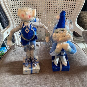 Puede incluir: Dos figuras caprichosas de duendes navideños. Un duende está de pie sobre una caja de regalo, vistiendo un atuendo azul y plateado con un diseño de copo de nieve. El otro duende está arrodillado sobre una caja de regalo, con un sombrero y atuendo azules.