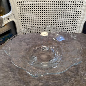 May include: A clear glass bowl featuring a scalloped edge and an etched floral pattern. The bowl has a shallow, wide basin with a smooth interior and a small white rectangular sticker attached to the inner rim near the top edge.
