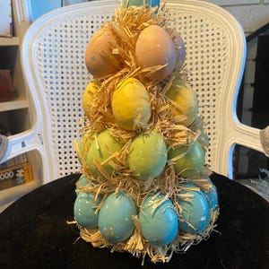 Puede incluir: Un árbol decorativo de huevos de Pascua con forma cónica. El árbol está compuesto por varios huevos de colores pastel, incluyendo azul, amarillo, verde y melocotón, anidados en paja. Los huevos están dispuestos en capas, creando una exhibición festiva y de temporada.