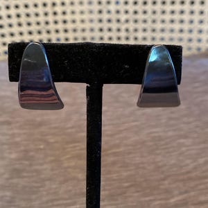 Könnte beinhalten: Ein Paar dunkelgraue, metallische, gebogene Creolen mit einer glatten, reflektierenden Oberfläche. Die Ohrringe haben die Form eines breiten, spitz zulaufenden Bogens und sind auf einem schwarzen Ständer präsentiert, der ihre polierte Oberfläche betont.