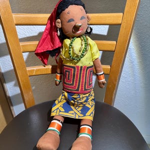 May include: A handcrafted doll wearing a red headscarf, yellow top, and patterned skirt. The doll is adorned with beaded necklaces, bracelets, and earrings. It sits on a wooden chair with a black seat. The doll's skin is brown.
