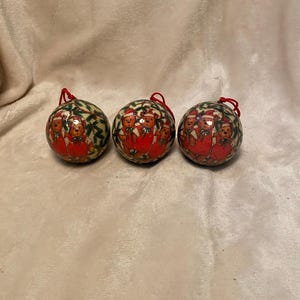 Op de afbeelding: Drie ronde kerstballen met rode ophangtouwtjes. Elke bal heeft een feestelijk ontwerp van teddyberen in rode outfits en kerstmutsen. De achtergrond van de ballen is een groen en goud patroon.