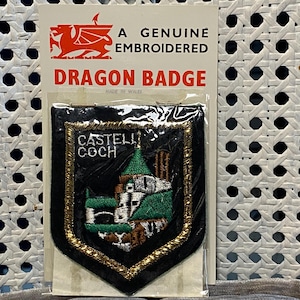 May include: A genuine embroidered Dragon Badge featuring a shield-shaped patch with a castle design in green, white, and brown, framed by gold trim. The badge is on a white card with a red dragon emblem and the text "DRAGON BADGE".