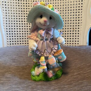 May include: A white bunny figurine wearing a light blue hat adorned with flowers and a lavender dress. The bunny holds two baskets and two Easter eggs. The figurine is standing on a patch of green grass.