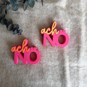 Könnte beinhalten: Zwei pinkfarbene Dekorationsobjekte mit den Worten "ach NO" in einer verspielten Schriftart. Das Wort "ach" ist in gelber Schrift, und "NO" ist fett und ohne Serifen. Die Objekte liegen auf einer neutralen, strukturierten Oberfläche.