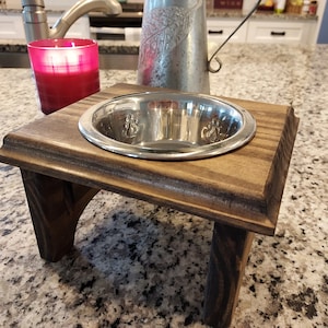 May include: A wooden pet food bowl stand with a stainless steel bowl. The bowl has paw prints embossed on the side. The stand is stained a dark brown color.