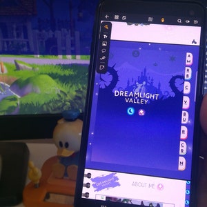 以下が含まれることがあります： 紫色の背景に白い城と月と星のアイコンが表示された、Dreamlight Valleyゲームをプレイしている携帯電話のスクリーンショットです。ゲームのタイトルは白い文字で表示されています。電話は机の上にあり、手で持たれています。